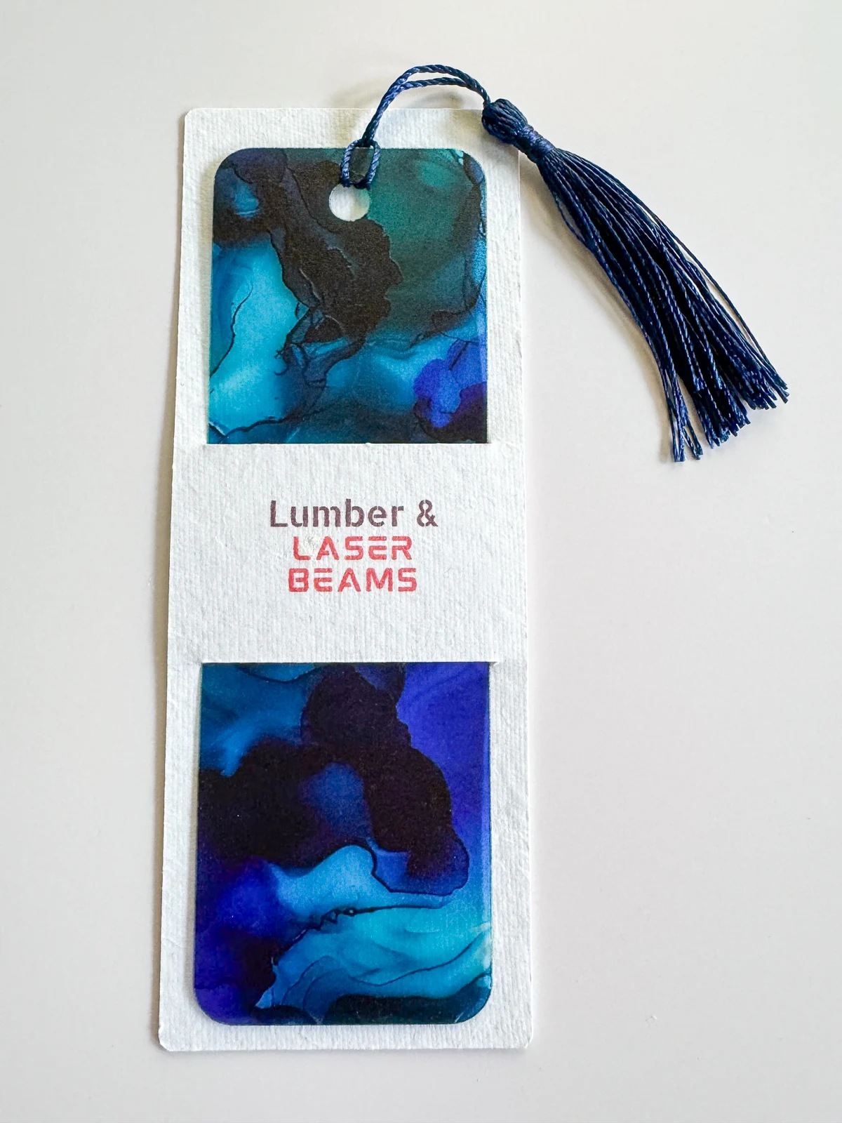 dark blue watercolor acrylic bookmark with dark blue tassel in a textured paper sleeve with the logo for the Etsy shop Lumber & Laser Beams on the front